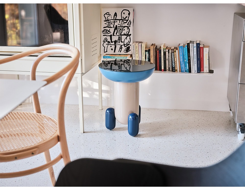 A playful side table with a glossy black top and a bold blue rim, supported by a cream-colored base and rounded legs. Its sculptural design brings a touch of whimsy to the reading nook.
