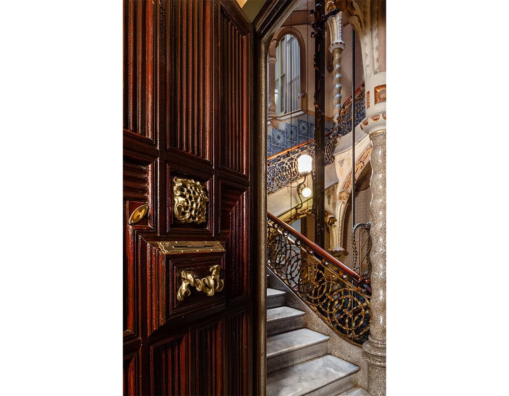 A polished brass Gaudí door knob is set into a carved wooden door. Behind it, a marble staircase and decorative iron railing complete the scene.