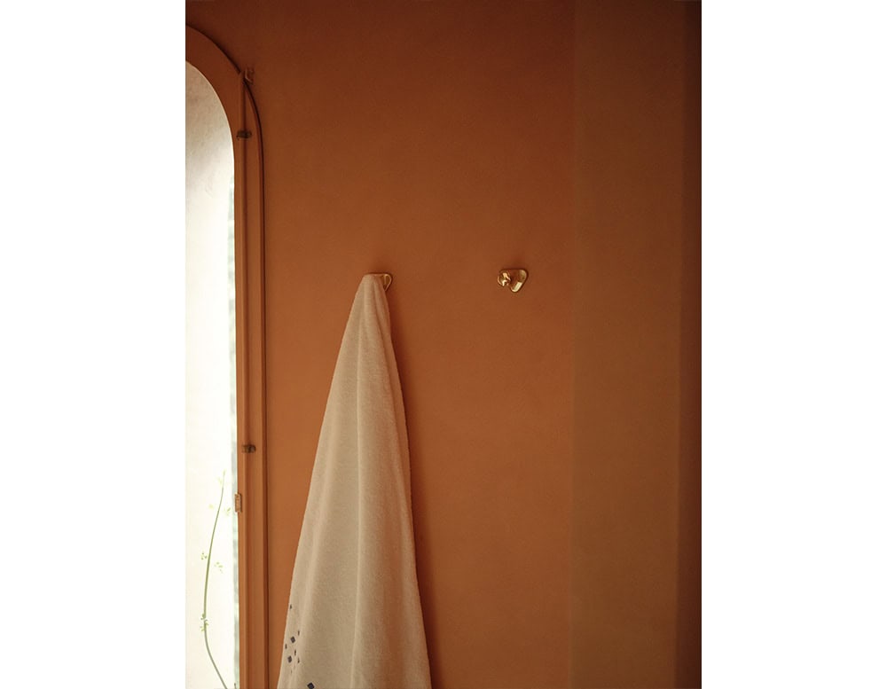 A pair of BD Gaudí Door Knobs are mounted on a terracotta-toned wall, their polished brass surfaces reflecting light. The simple setting is complemented by an arched window and a soft towel casually hanging from one of the knobs.