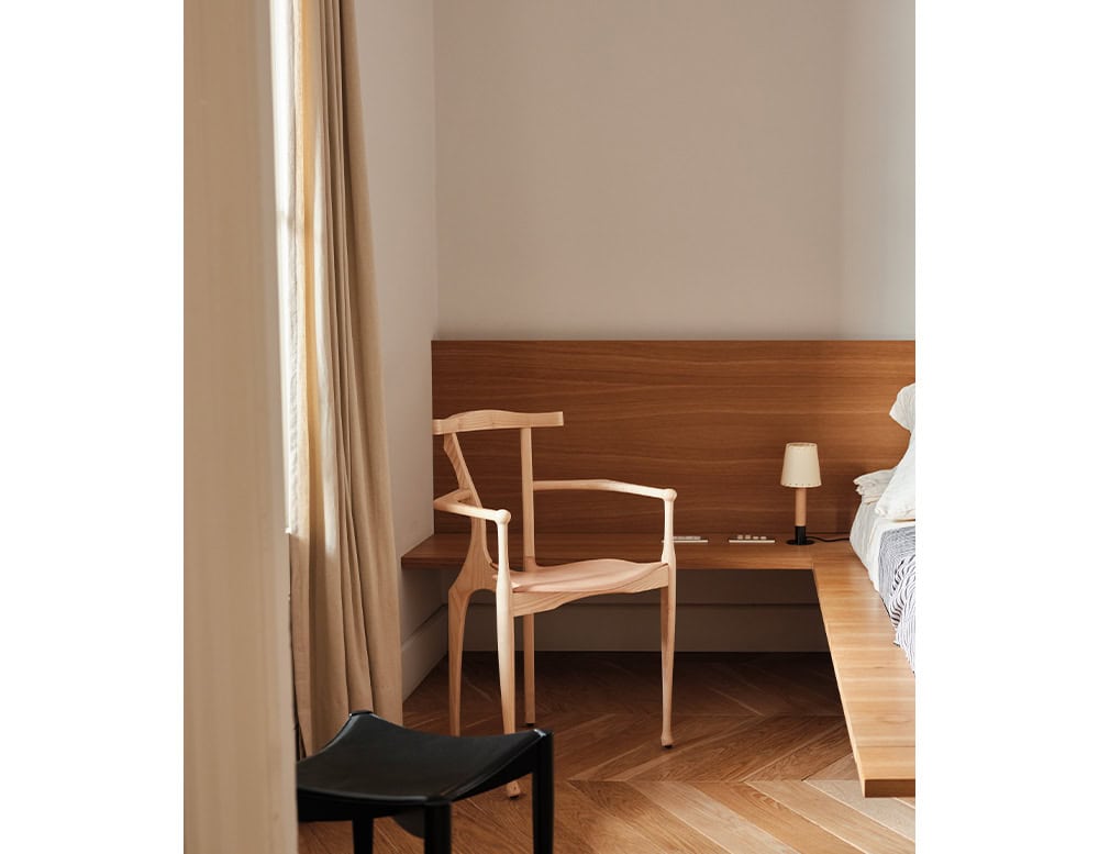 A pale wood BD Gaulino Chair is placed beside a low wooden platform bed. Its sculptural frame complements the soft, minimal bedroom setting.