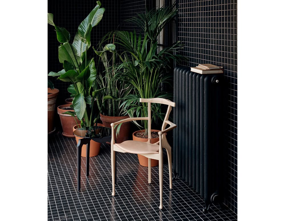 A light cream BD Gaulino Chair sits among potted plants on a black tiled floor. Its sculptural form contrasts softly with the grid backdrop and greenery.