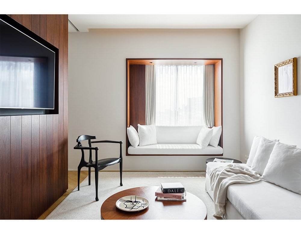 Bright, minimalist living space featuring the sculptural Gaulino Easy Chair in black, paired with a built-in wood-framed window seat, white sofas, and a round coffee table. Warm wood tones contrast elegantly with soft neutrals and natural light.