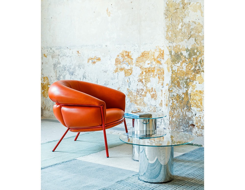 The BD Grasso Armchair in orange leather is shown against a textured wall. The chair has padded, overstuffed arms and a rounded backrest, supported by a red metal frame. It is paired with two glass and chrome tables.