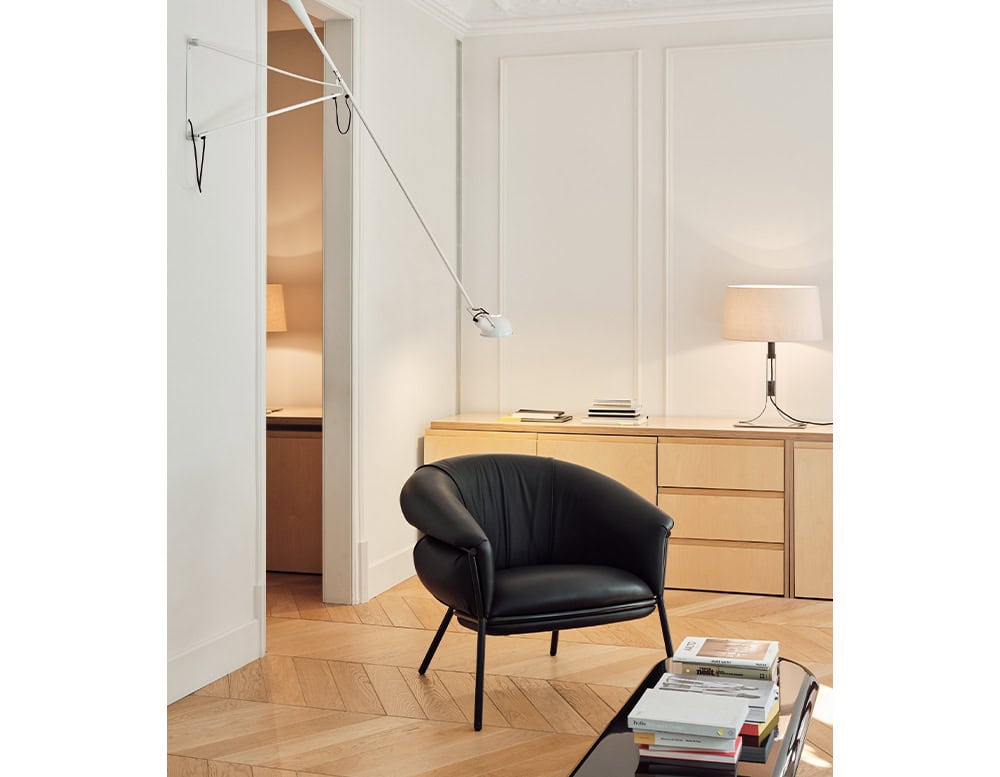 The BD Grasso Armchair is shown in a minimalist interior. The chair features black leather upholstery with rounded, padded arms and a metal base. It is positioned next to light wood cabinetry and a modern reading lamp.