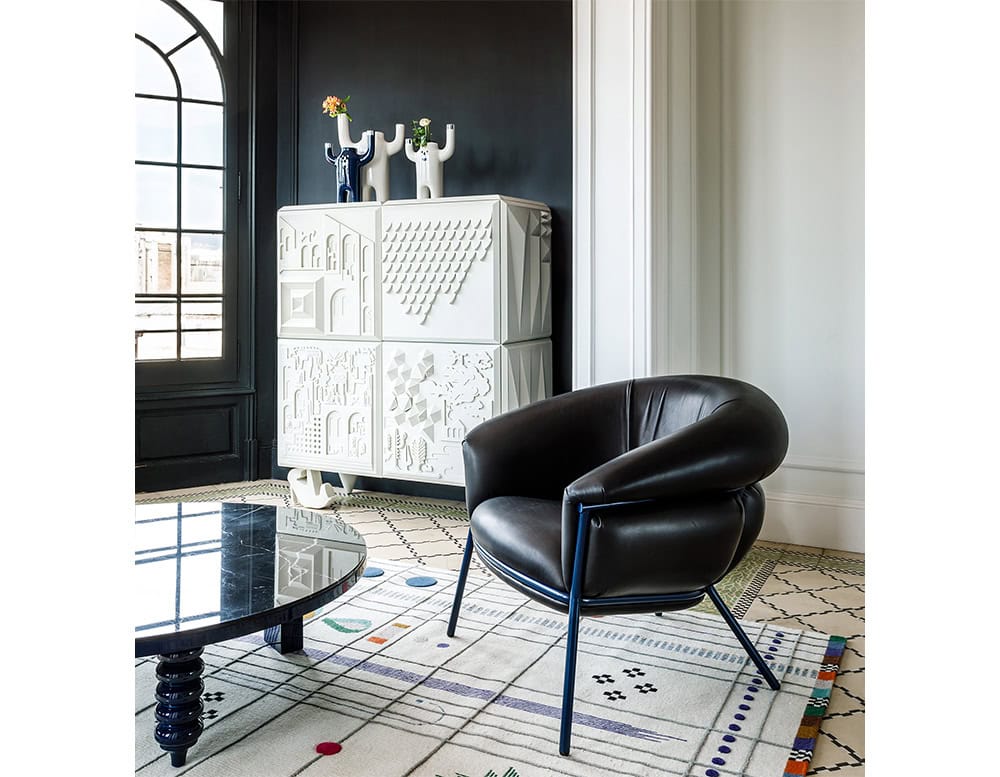 Ceramic faces sit atop a sculptural white cabinet with intricate geometric reliefs. A sleek black leather armchair, patterned rug, and round black coffee table complete the eclectic, design-forward interior with a mix of whimsy and sophistication.