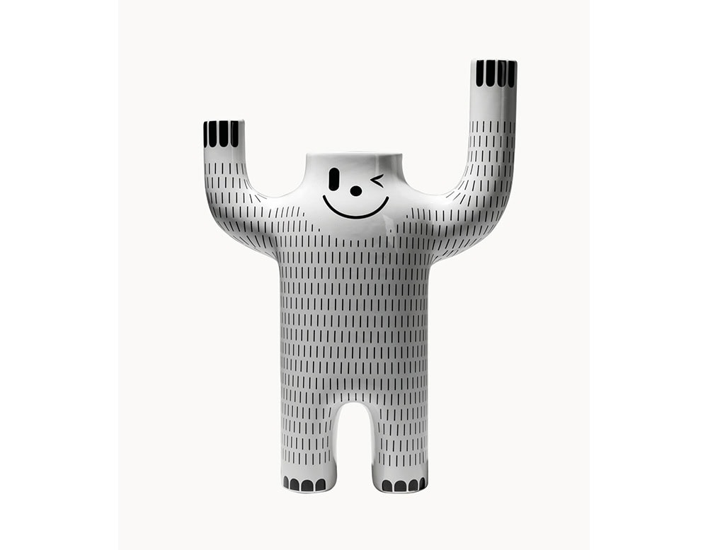 White ceramic figure with raised arms, a winking smiley face, and a vertical line pattern across its body. Sculptural and fun, with a bold, character-like presence.