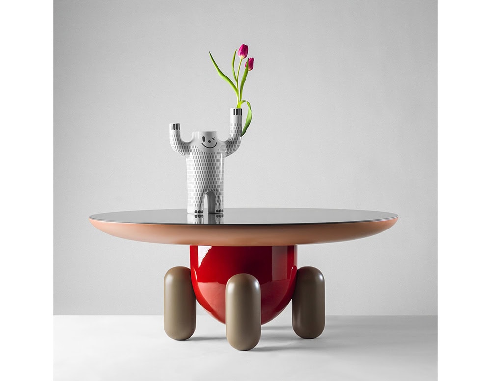 Joyful figure and raised arms. It sits atop a bold, sculptural table featuring a glossy red base, rounded legs, and a sleek circular top.
