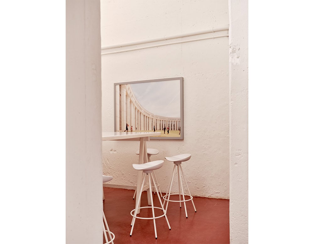 Compact space with four white Jamaica Stools around a tall table, set against textured white walls and red flooring. A framed architectural photo adds visual depth and elegance to the minimalist corner.
