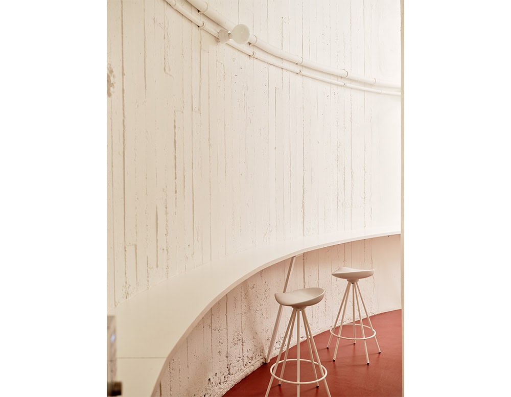 Curved white bar with two Jamaica Stools set against a textured, vertical-lined wall. The soft tones and minimalist shapes contrast with the red flooring, creating a calm, sculptural atmosphere.