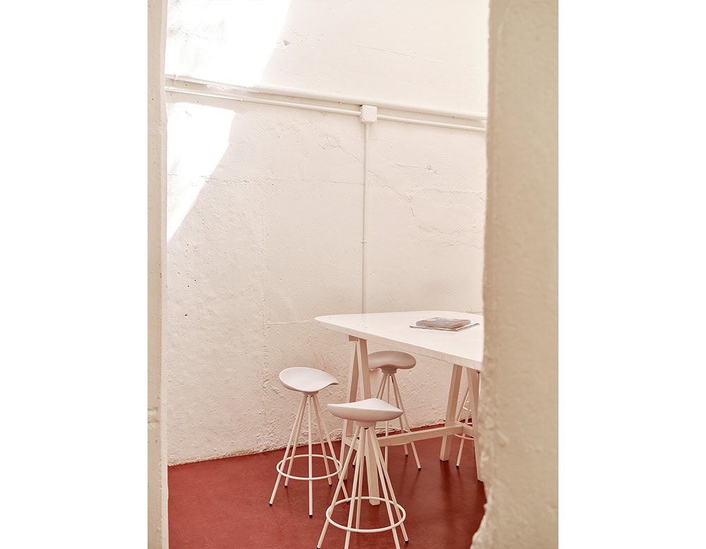 Minimalist corner setup with white Jamaica Stools and a matching high table, all set against textured white walls and a bold red floor. The space feels airy, clean, and quietly sculptural.