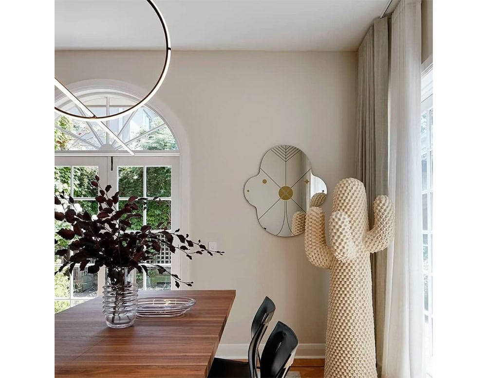 Modern dining area featuring a sculptural cactus coat rack, floral wall mirror, round LED pendant, and wood table with a ribbed glass vase. Soft curtains and arched windows complete the bright, artistic space.