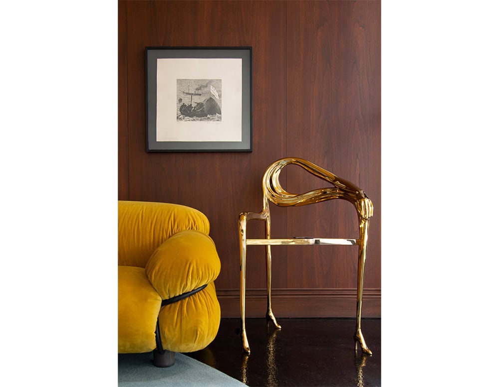A polished bronze armchair with surreal, organic lines sits beside a mustard velvet lounge chair. The composition highlights a bold mix of textures and finishes against a warm wood-paneled wall.