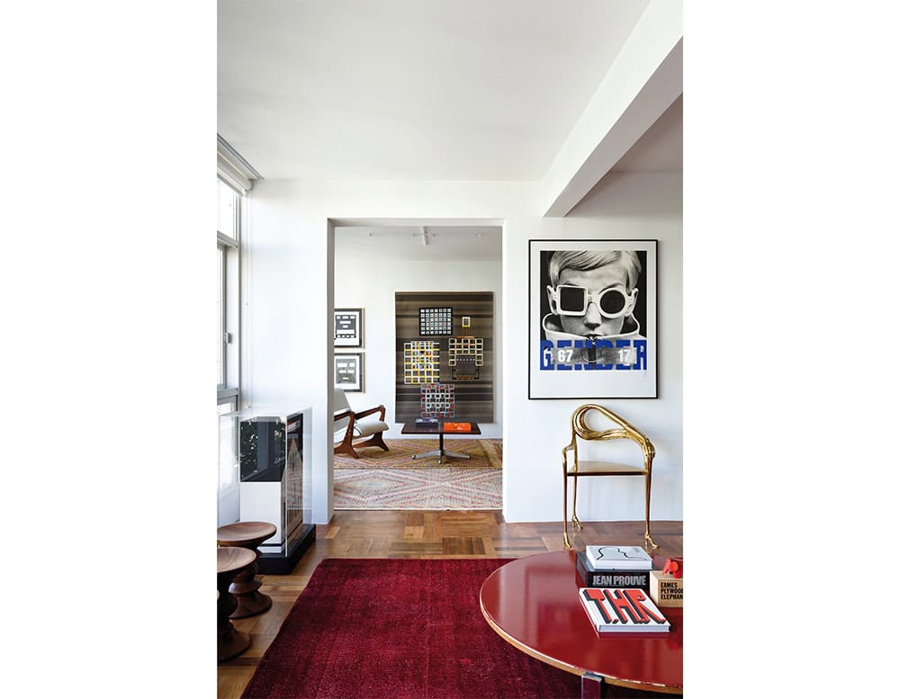 A polished bronze armchair is placed beside a red lacquered coffee table, adding a sculptural note to a living room layered with modern art, geometric furniture, and bold graphic prints.