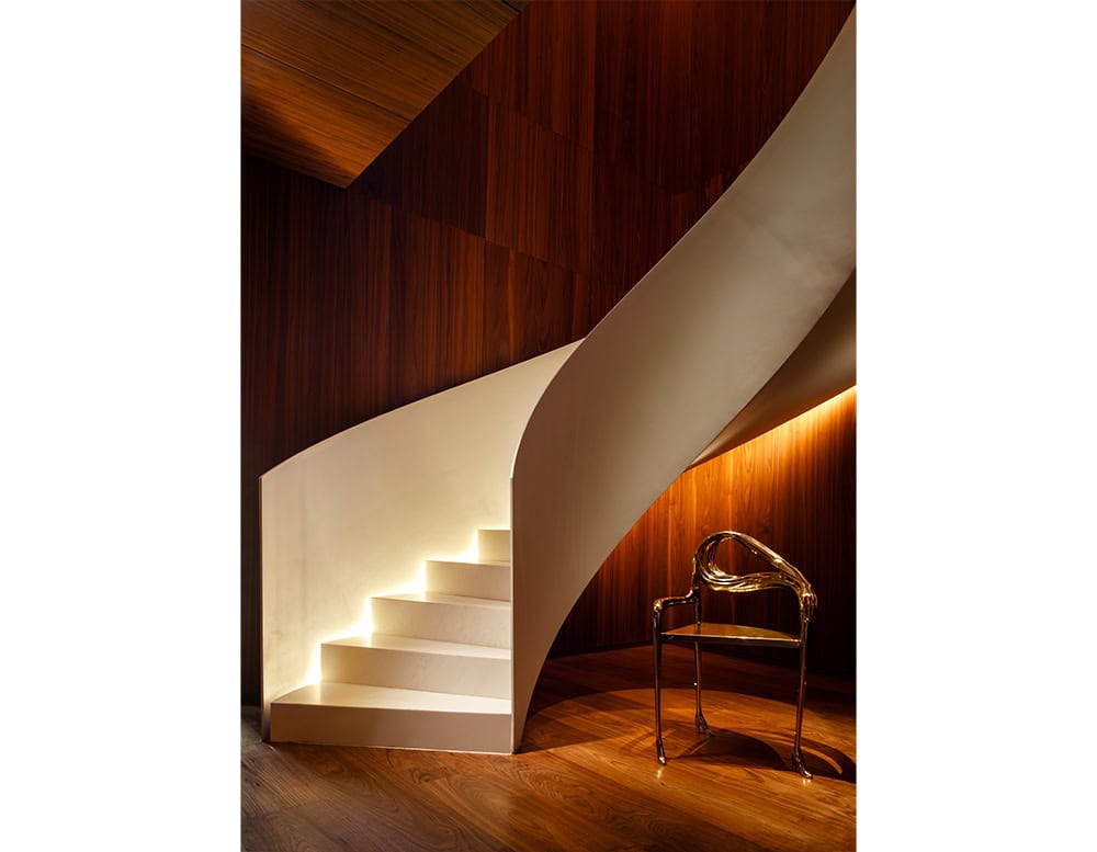 A sculptural armchair in polished bronze sits at the base of a softly lit spiral staircase. Its flowing, organic lines echo the curves of the architecture, creating a dialogue between furniture and space.