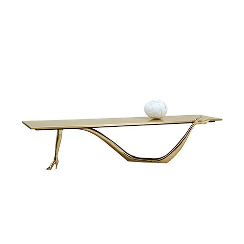 A bold coffee table with a golden tabletop supported by sculptural bronze legs. One leg mimics a high-heeled foot while the other is a graceful arm, creating a whimsical and surreal silhouette. A marble sphere rests on top.