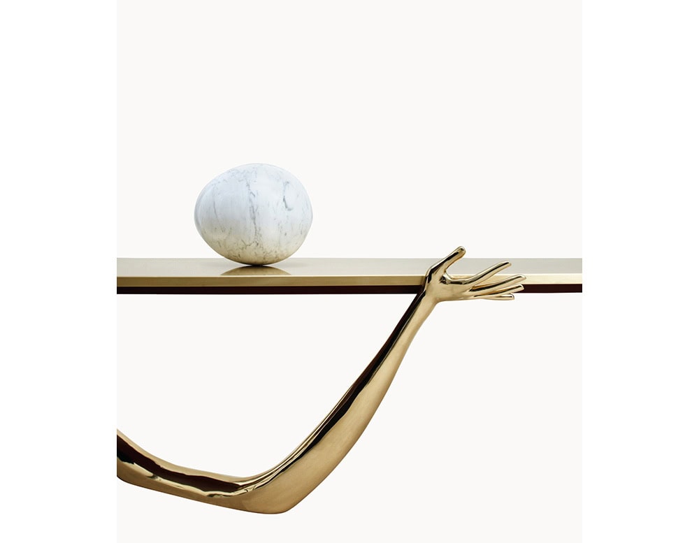 A surreal coffee table featuring a polished bronze arm gracefully supporting the golden top. A marble sphere rests on the surface, adding an unexpected sculptural accent to the piece.