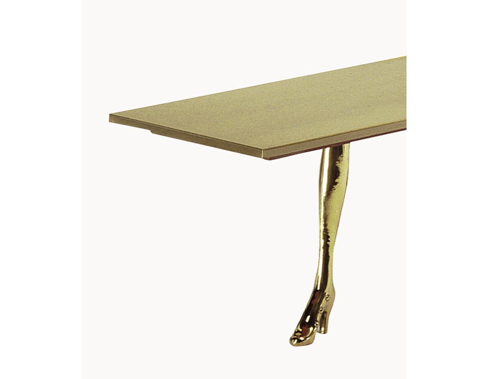 A sculptural coffee table featuring a sleek, rectangular top in brushed golden tones. The base surprises with a surrealist detail, a polished cast-bronze leg in the shape of a woman’s leg and foot, wearing a delicate high heel.