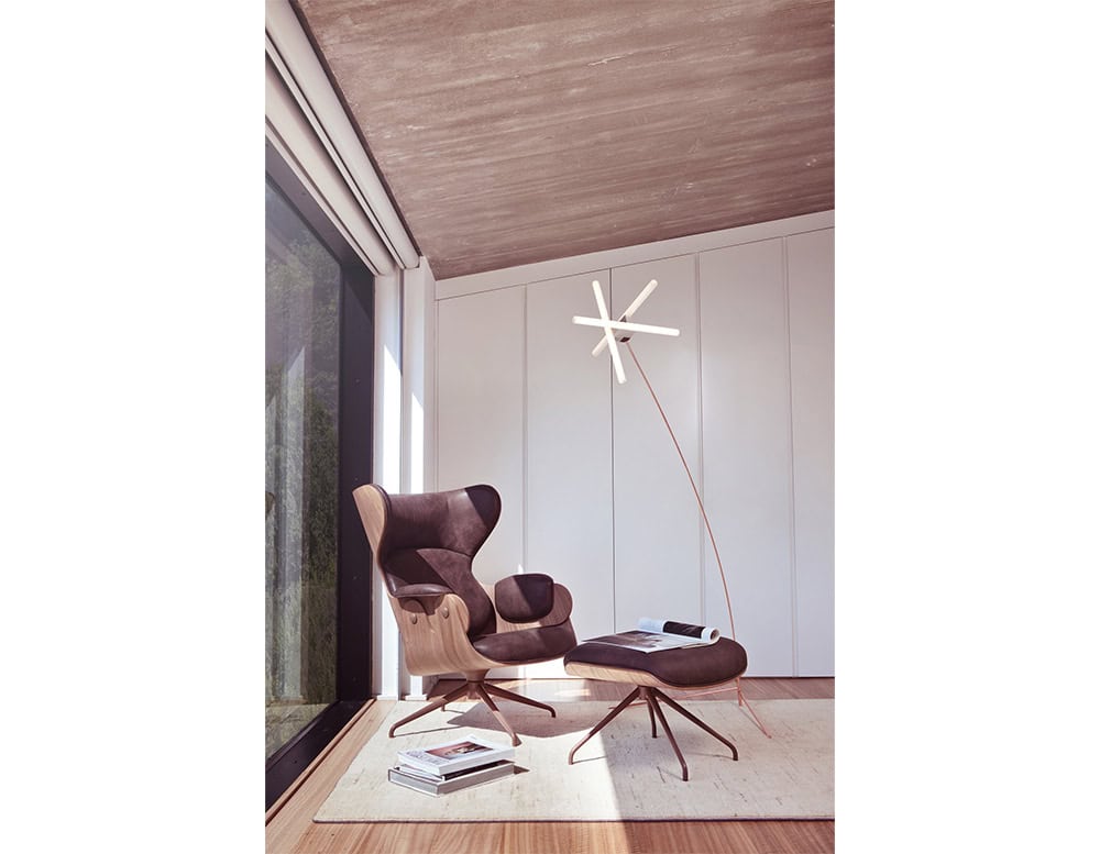 Stylish lounge corner with a Lounger Armchair and matching ottoman in dark upholstery and curved wood shell. Positioned beside a floor-to-ceiling window and accented by a sculptural floor lamp, the space is calm, modern, and perfectly lit.