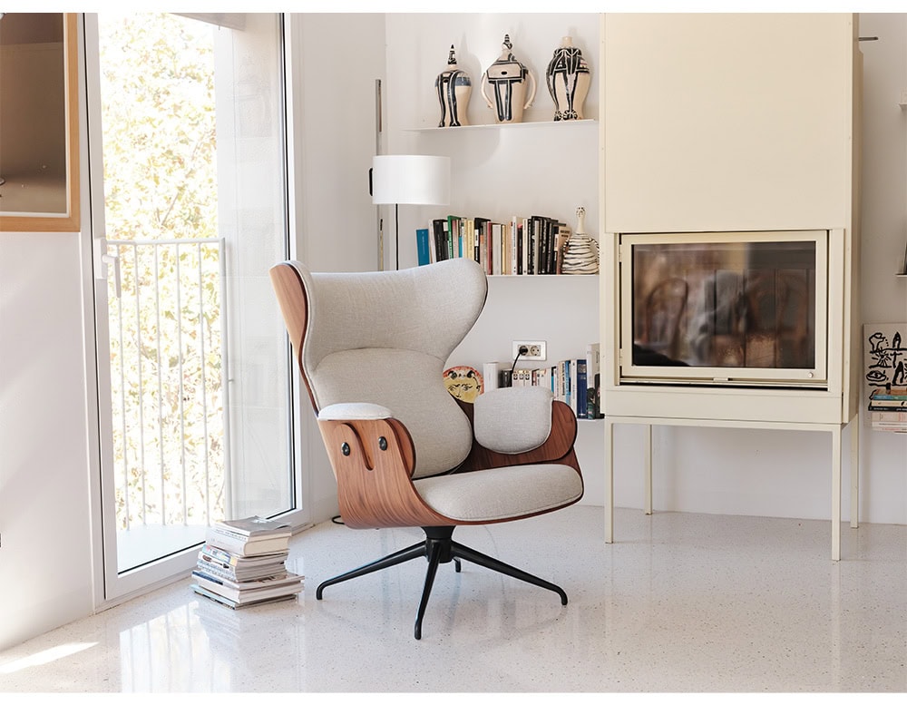 Contemporary reading nook with a Lounger Armchair featuring a wood shell and light gray upholstery. Surrounded by books, ceramics, and natural light, the space feels warm, artistic, and inviting.