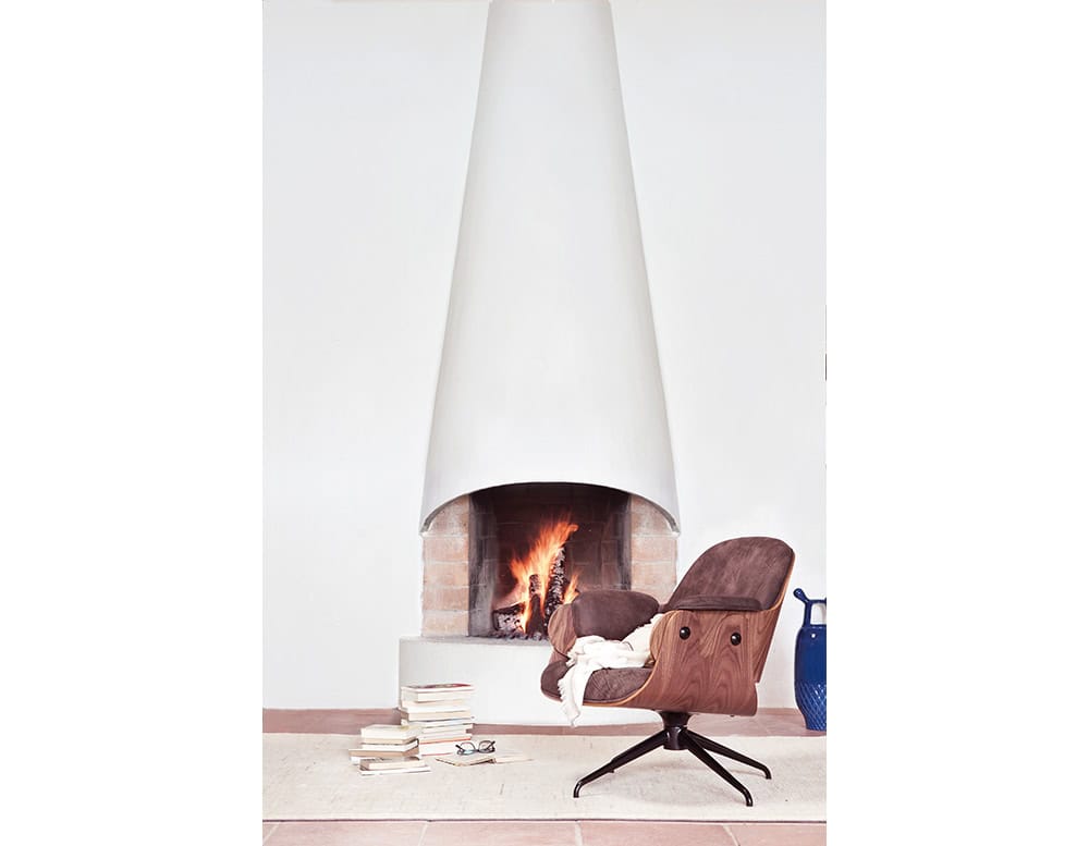 Low Lounger Armchair in warm suede and walnut shell. Set by a lit conical fireplace, beside stacked books and a cobalt ceramic vase, creating a cozy reading nook with soft light and texture contrast.