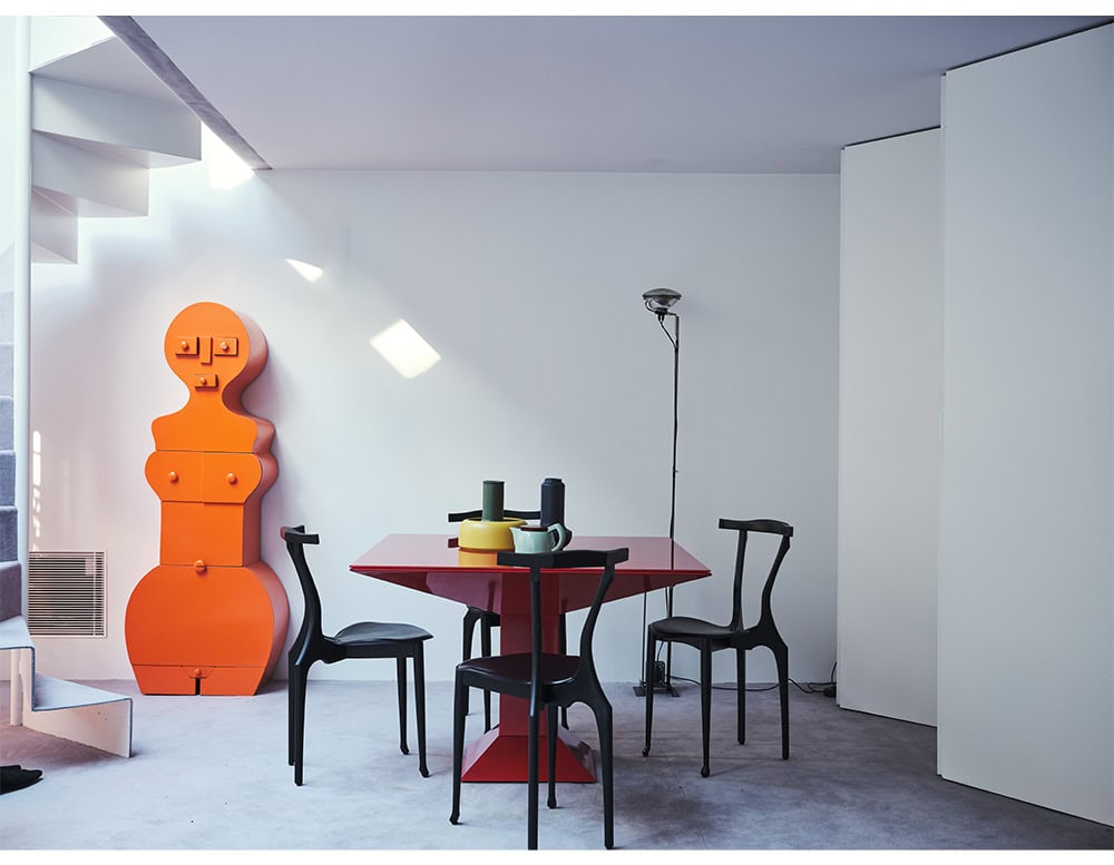 Mettsass Table in glossy red with sculptural base, paired with matte black chairs and colorful ceramic vases. Scene includes an orange totem cabinet, minimalist stair, floor lamp and soft lavender carpet in a modern white interior.