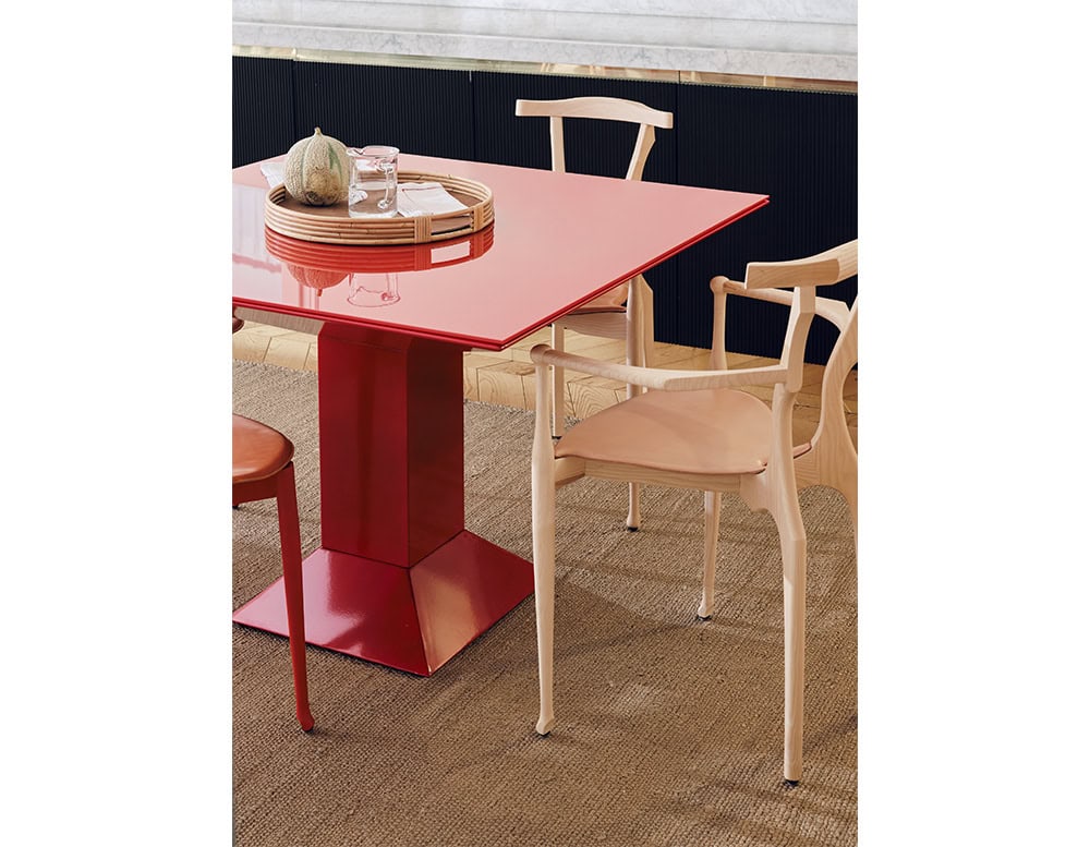 Mettsass Table in glossy red finish with a bold geometric base. Styled with a natural woven tray, glassware, a melon centerpiece, and surrounded by pale wood dining chairs over a textured tan rug and parquet floor.