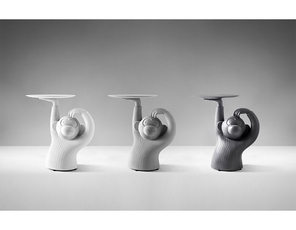 Three monkey-shaped side tables in soft gradient tones from light to dark grey. Each piece features smooth, rounded lines with one arm playfully holding up a circular tabletop. The textured surface highlights the sculptural detail.