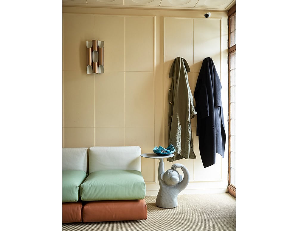 Monkey-shaped side table with a raised tray, placed by a two-tone modular sofa. The tabletop holds a textured ceramic piece, while coats hang casually on the wall nearby.