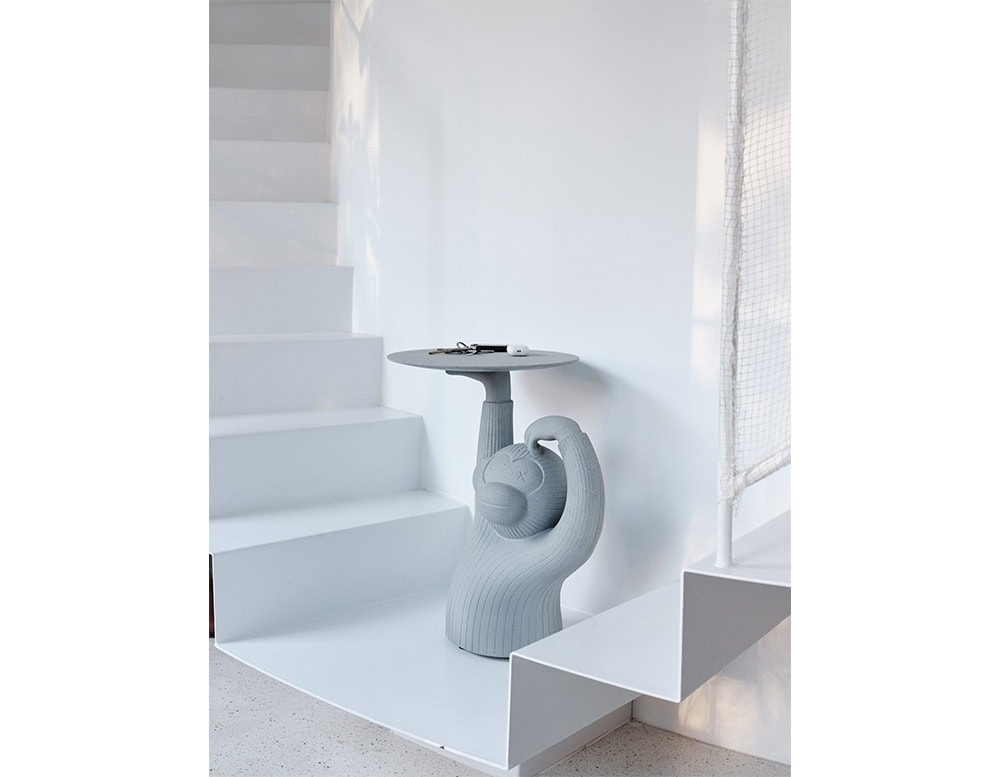 A playful side table featuring a monkey figure supporting a round tabletop, placed next to a white staircase with keys and a small device resting on the surface.