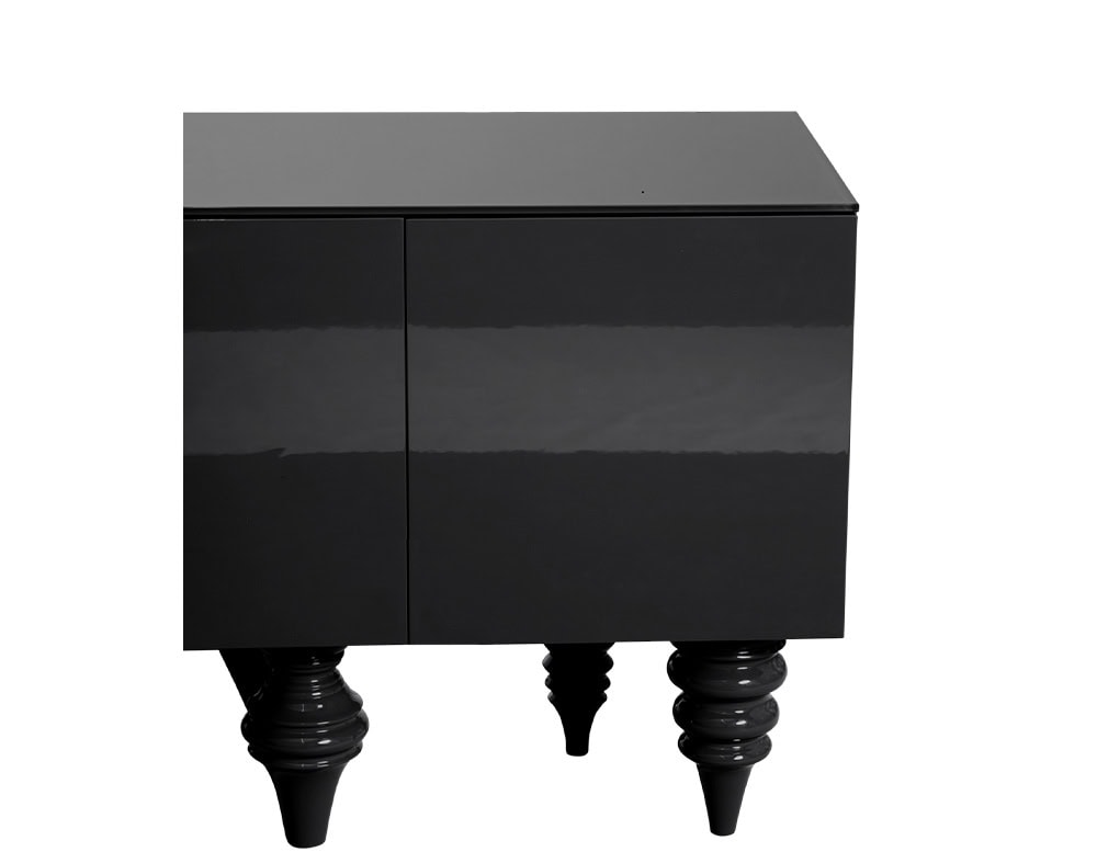 A deep black cabinet with a glossy polished finish. The front is sleek and understated, while the base stands on a composition of turned and sculptural legs that introduce playful contrast to the cabinet's clean lines.