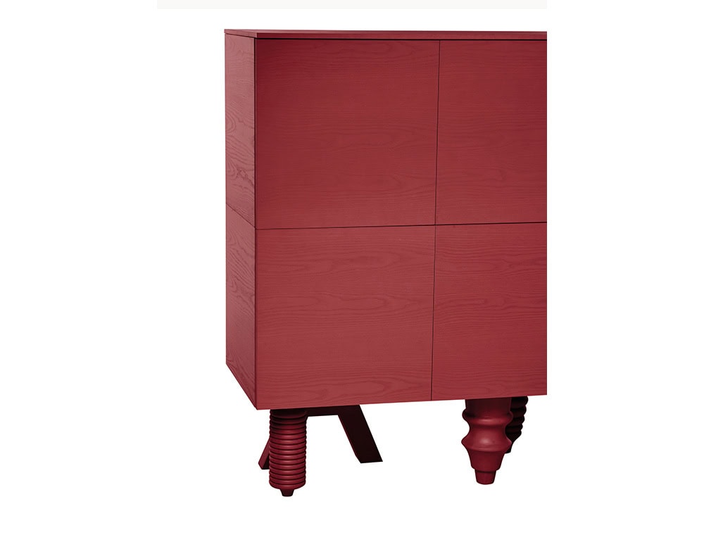 Crimson-toned cabinet with clean vertical panels, raised on sculptural supports in playful silhouettes, blending symmetry with surprise in a compact, upright form.