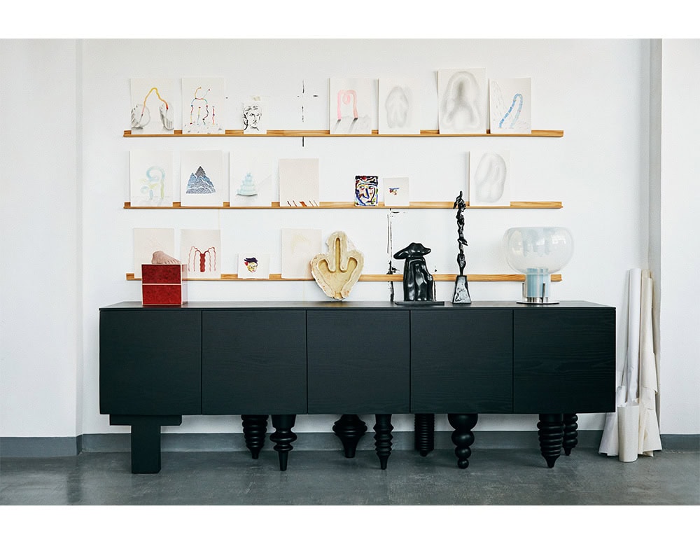 Black wood cabinet with unique sculptural legs. Topped with eclectic decor like a ceramic hand, black sculptures, and a glass lamp. Framed by a wall display of artworks and wooden rails, with rolled textiles placed casually at the side.