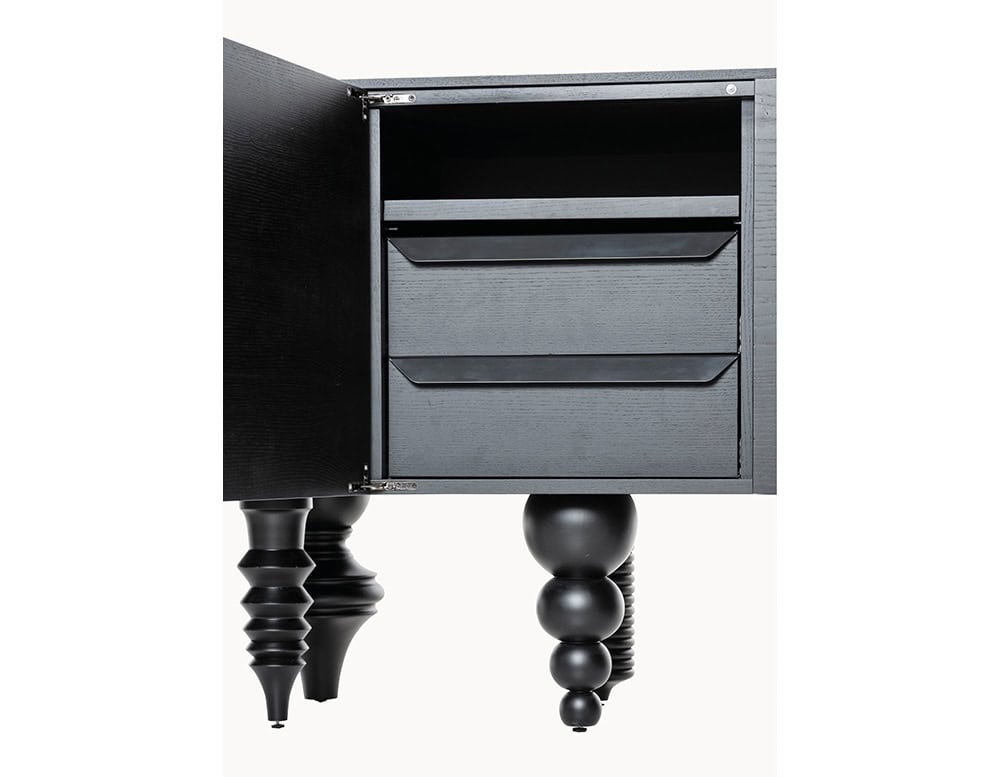Black wood cabinet open to reveal interior drawers and shelves. Features sculptural turned legs in varied shapes. The focus is on craftsmanship and playful geometry, with high contrast between the matte black finish and crisp white background.