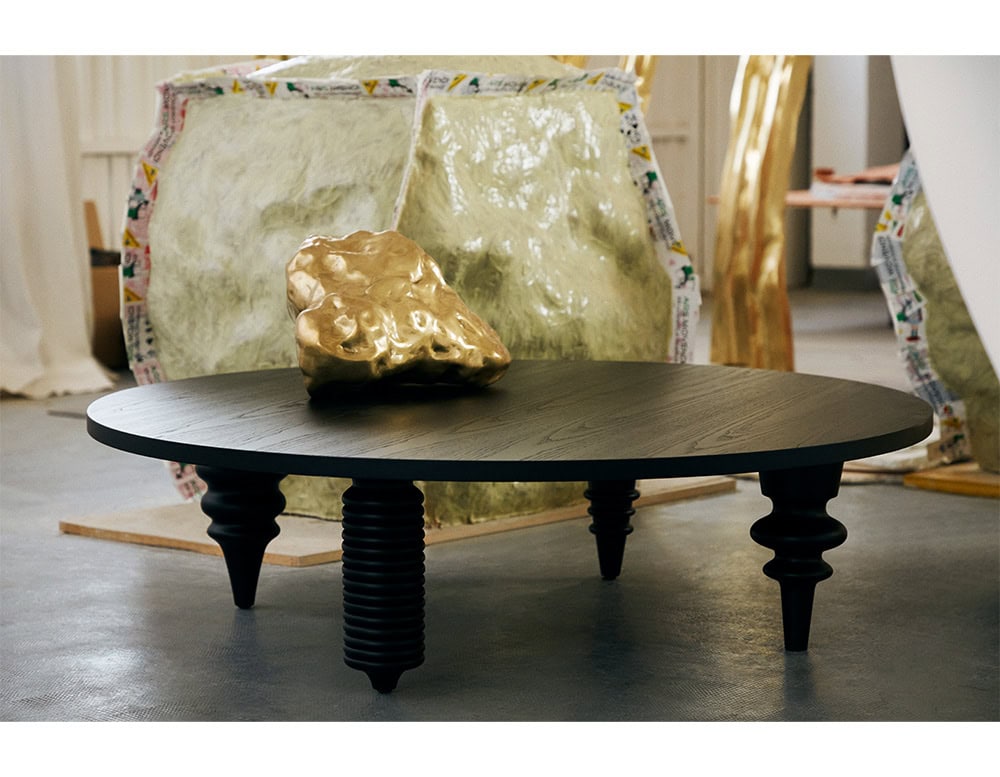 Multileg Coffee Table in matte black with turned wood legs in varying profiles, set against sculptural forms and a textured gold object, blending art and function.