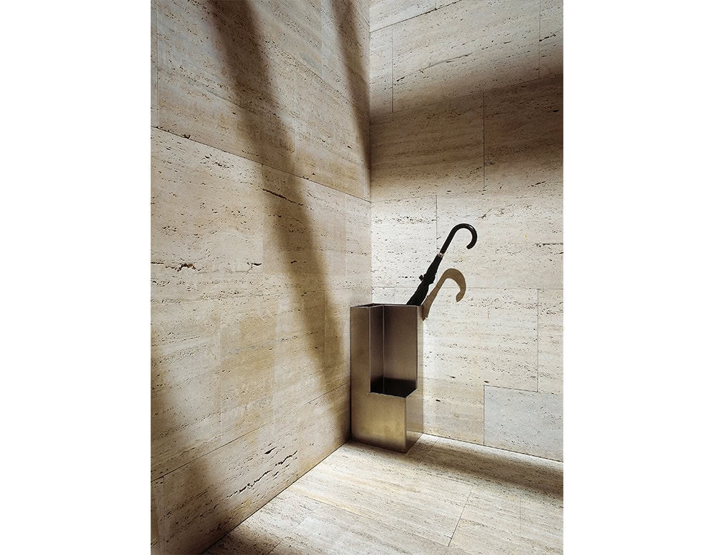 A polished metal umbrella stand with architectural folds, placed in a minimalist travertine-lined corner, holding a single classic black umbrella that casts a dramatic shadow on the wall.