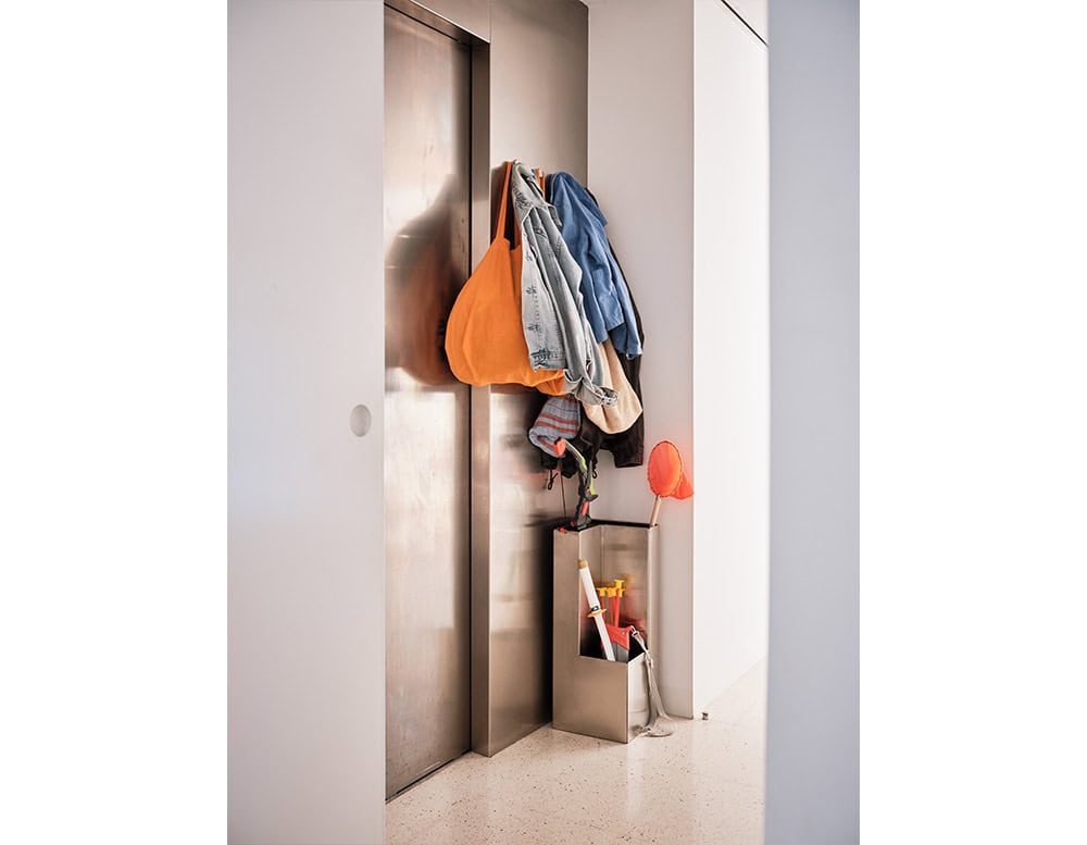 Tall and slim metal coat rack and umbrella stand in a satin finish, holding casual outerwear, bags, and assorted everyday accessories in a functional, minimal entryway scene.
