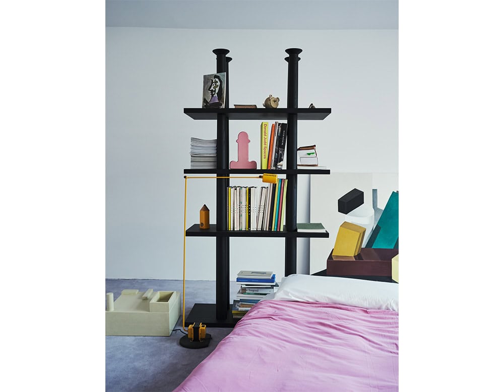 A pink ceramic vase is displayed on a black open-shelf bookcase, surrounded by books, objects, and graphic art. A yellow floor lamp adds a bold accent near a bed dressed in soft lavender bedding.