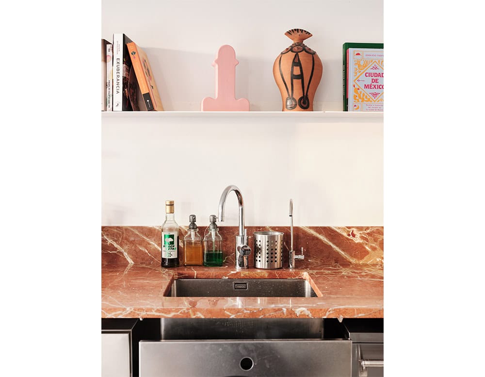 A pink ceramic vase adds a playful touch to the kitchen shelf, paired with books and a terracotta vessel. Below, a red marble countertop frames the sink.