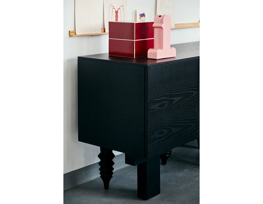 A pink ceramic vase with bold, sculptural lines sits atop a dark wood cabinet with carved, playful legs. Next to it, a red lacquered box adds a contrasting pop of color, while delicate sketches line the wall above.