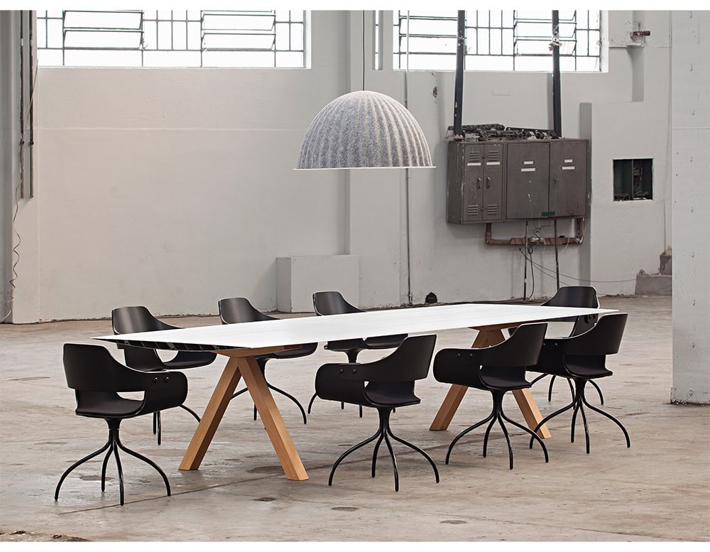 The BD Table B is placed in an open industrial space, combining a white aluminum tabletop with natural wooden legs. The setting contrasts raw architectural elements with the table's clean, refined lines.