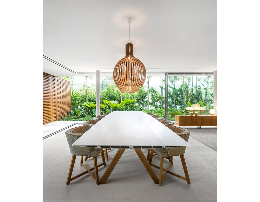 The BD Table B is shown in a contemporary dining space, featuring a sleek white tabletop and warm wooden trestle legs. The surrounding environment highlights natural elements, with lush greenery seen through expansive glass walls.