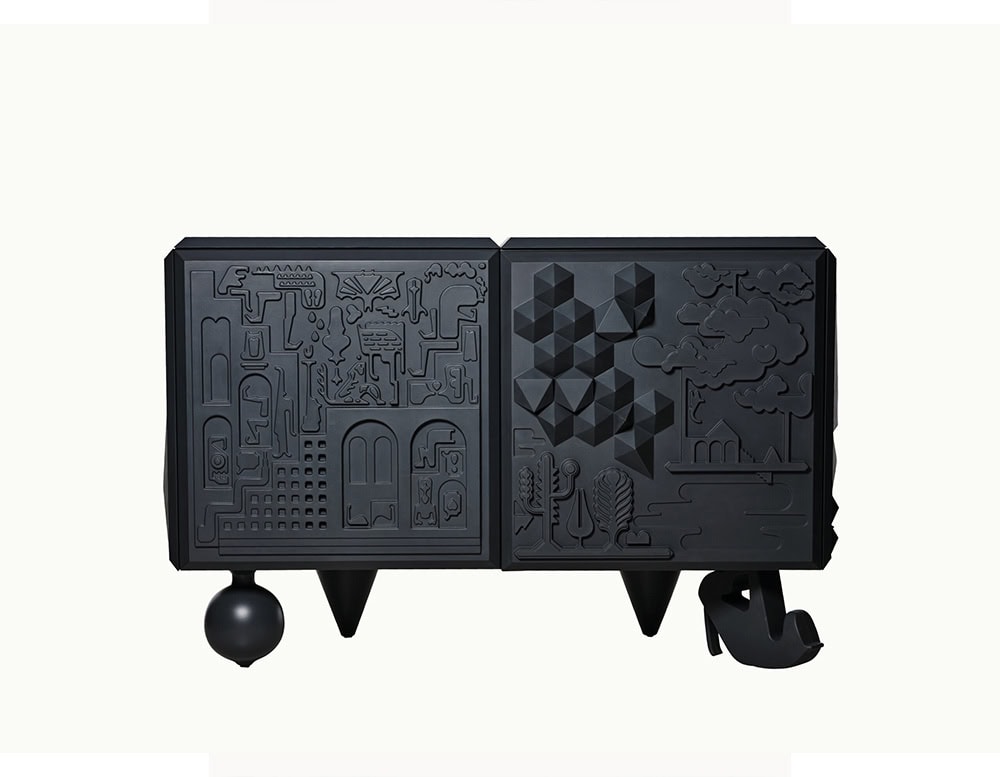 Matte black cabinet with two intricately carved doors displaying architectural and geometric reliefs. The base combines a sphere, tapered cone, and a whimsical shoe-shaped foot for an eclectic, sculptural profile.