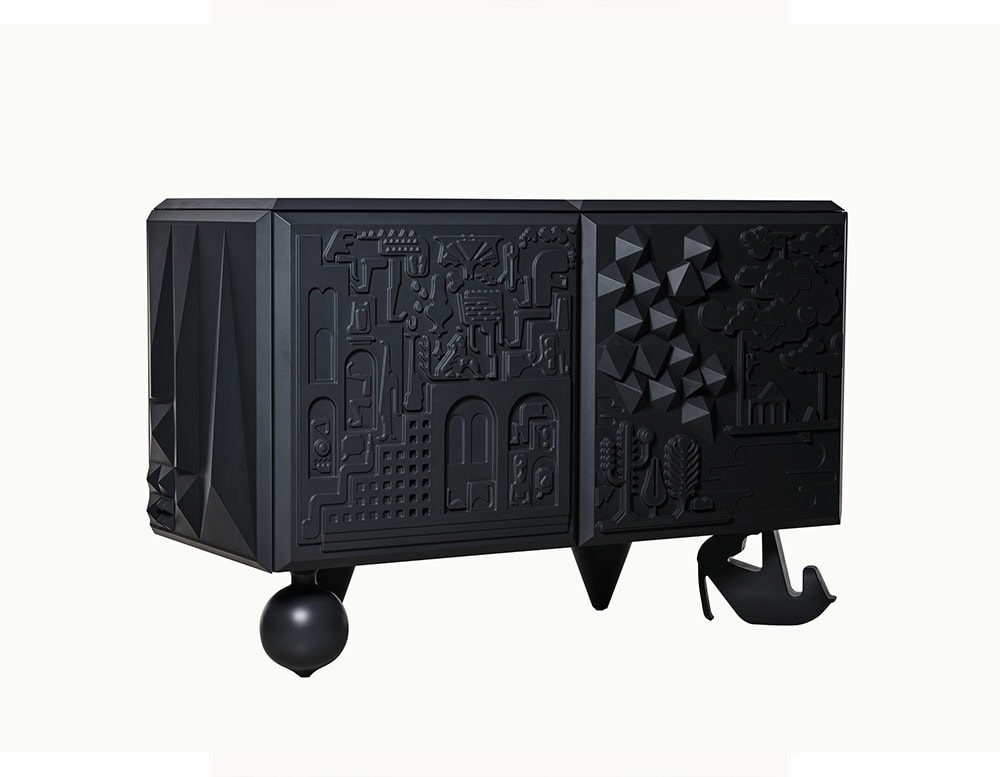 Black cabinet with sculpted relief doors featuring abstract scenes, resting on playful asymmetrical legs, including a spherical and shoe-shaped base, with sharply faceted textures along the side.