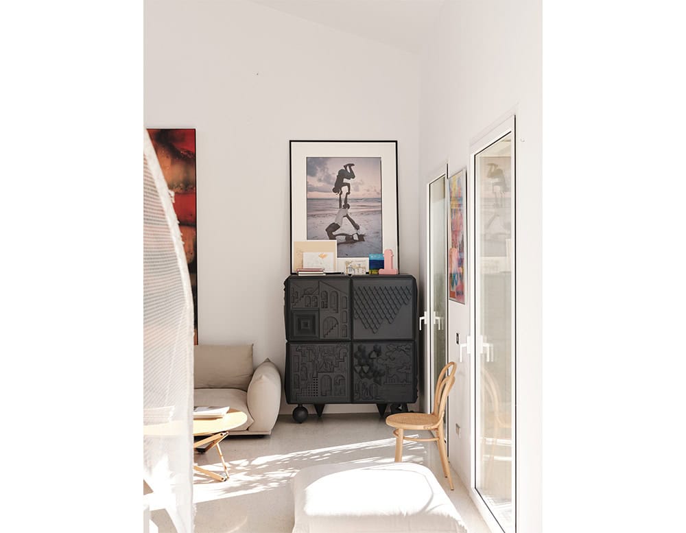 Compact black cabinet with textured, sculptural doors, placed under a framed photograph and decorated with books and objects. Surrounded by a lounge chair, bentwood chair, and natural light through glass doors.