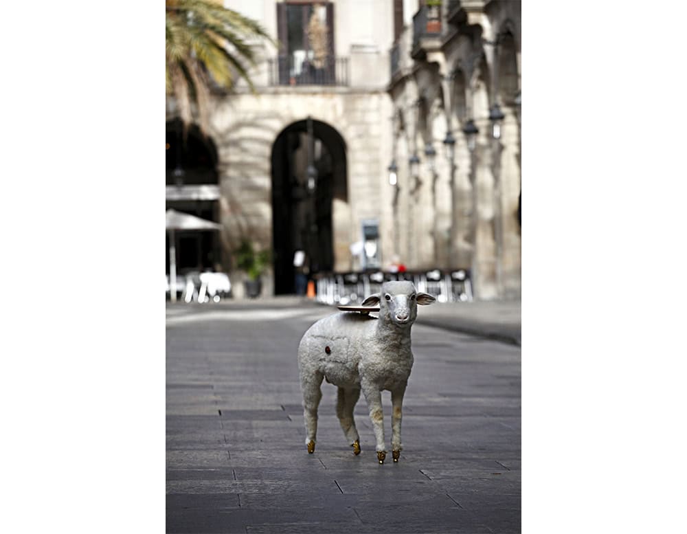 A sheep-shaped side table with a playful, surreal twist is seen standing in the middle of a historic plaza. Its soft fleece contrasts with polished brass hooves and a wooden tabletop.