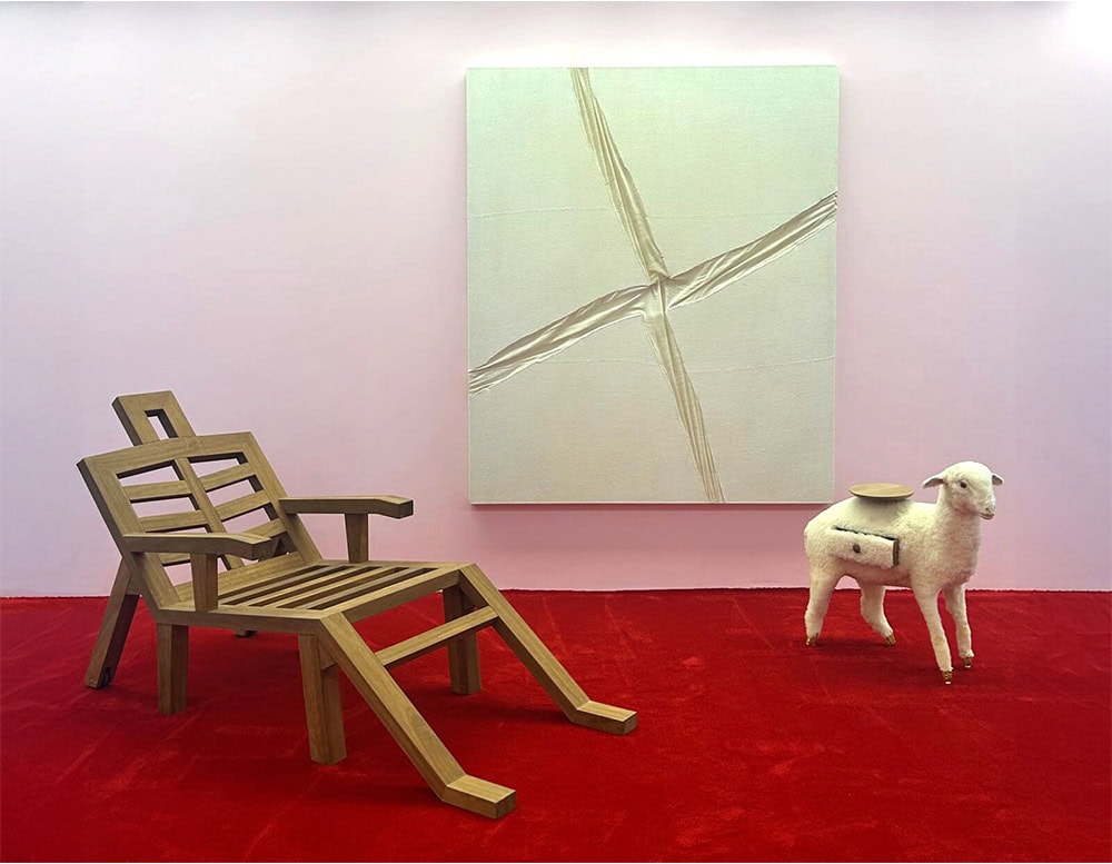 A sculptural wooden lounge chair with bold, angular lines and an exaggerated silhouette rests on a vivid red carpet. To the side, a whimsical lamb-shaped side table adds a playful touch, while a textured monochrome canvas completes the scene.