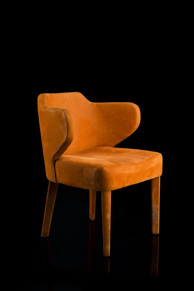 EX Tra Chair upholstered in orange leather and four wood legs on a black background.