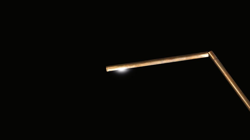 Closeup of Pipe Light-L: L-shaped bronze lamp with a bulb at the end and red wiring on a black background.