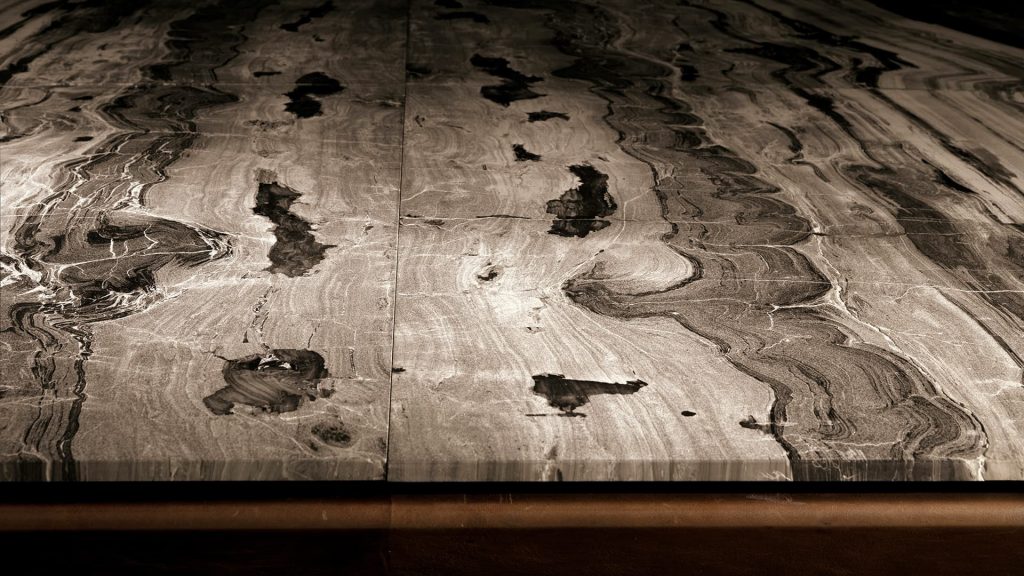 Closeup of Soprano dining table, structure and four legs in brown wood, top in white, grey, brown and black stone on a black background.