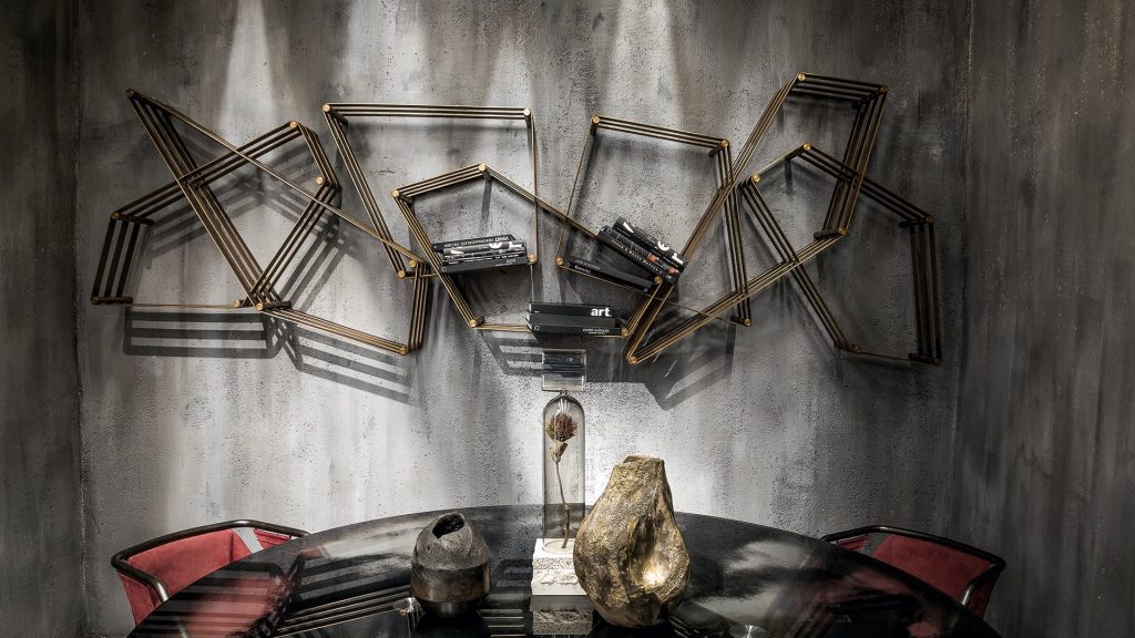 Tangram bookcase made of irregular polygons joined together. Structure in natural burnished brass attached to a gray wall in a dining room.